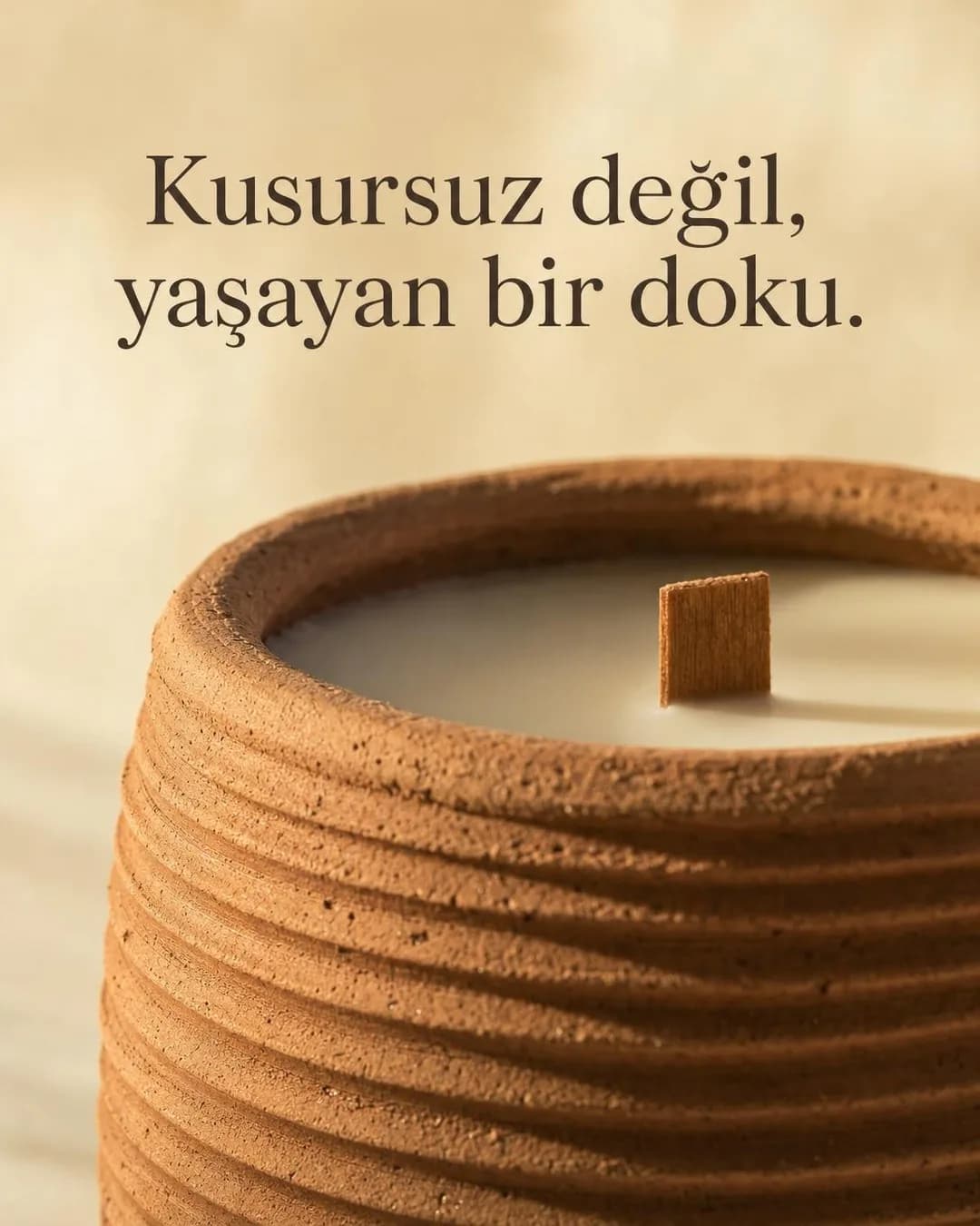Ham, pişmiş toprak tonlarında yatay dokulu sırsız seramik mum kabının yakın çekimi. Kabın içinde düzgün dökülmüş krem rengi mum ve dik duran ince dikdörtgen ahşap fitil görülüyor. Üstte koyu kahverengi serif fontla 'Kusursuz değil, yaşayan bir doku.' yazısı yer alıyor.