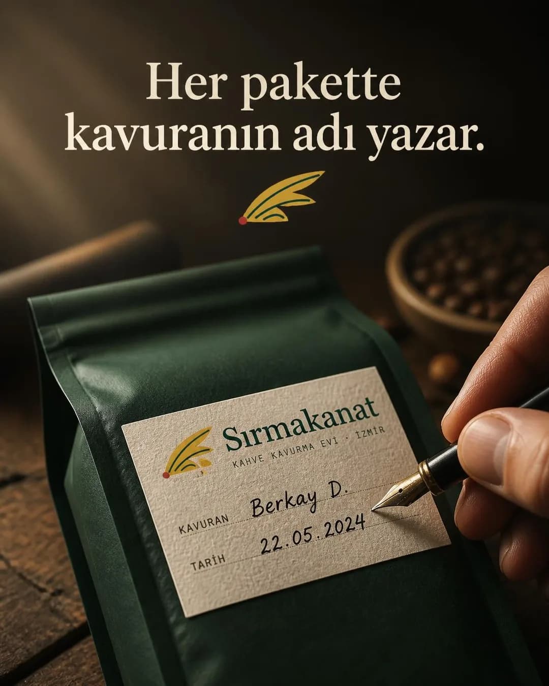 Koyu yeşil kahve paketinin üzerindeki krem rengi etikete dolma kalemle yazı yazan bir el. Etikette Sırmakanat logosu, el yazısıyla 'Berkay D.' ve '22.05.2024' tarihi görülüyor. Paketin üst kısmında zarif bir tırnaklı yazı tipiyle 'Her pakette kavuranın adı yazar.' başlığı yer alıyor; arka planda ahşap zemin ve kahve çekirdekleri hafif flulaşmış şekilde duruyor.