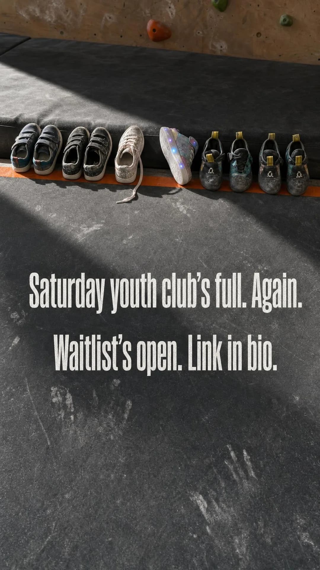 A row of kids' shoes — sneakers and tiny rental climbing shoes — kicked off at the edge of the foam matting, every toe pointing toward the bouldering wall. Text in the frame: Saturday youth club's full. Again. Waitlist's open. Link in bio.
