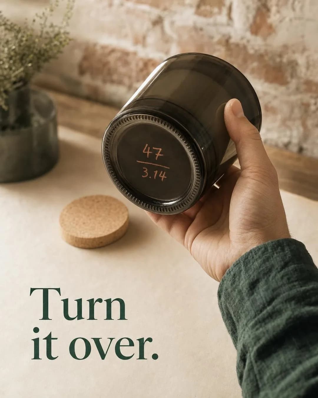 A hand in a dark green sleeve holds a dark smoked-glass candle jar tilted up to reveal handwritten terracotta-colored numbers on the base: 47 over 3.14. On the warm paper surface behind it rests a round cork lid and a small green sprig in front of a soft brick wall. Dark green serif text in the lower left reads, 'Turn it over.'