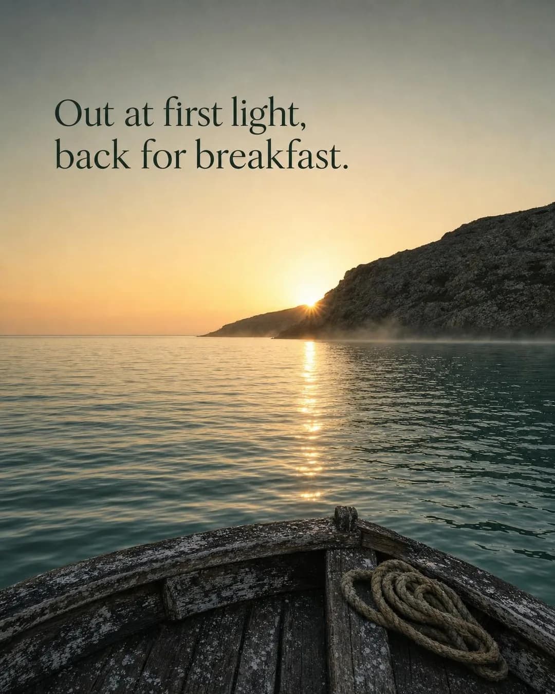 View from the weathered wooden bow of a small boat on calm, turquoise coastal water at sunrise. The sun peeks over the ridge of a dark, rocky cliff where a low band of mist gathers over the water. Warm golden sunlight reflects across the surface. At the upper left, text reads: 'Out at first light, back for breakfast.'
